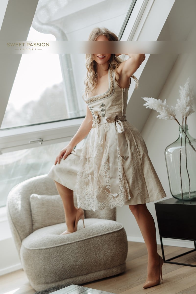 Laura high class escort muenchen steht, mit einem Bein, auf einem grauen Seßel. Sie trägt ein beiges Dirdl, ohne Bluse, welches aufwenidig verziert ist. Dazu trägt sie nudefarbene Highheels.